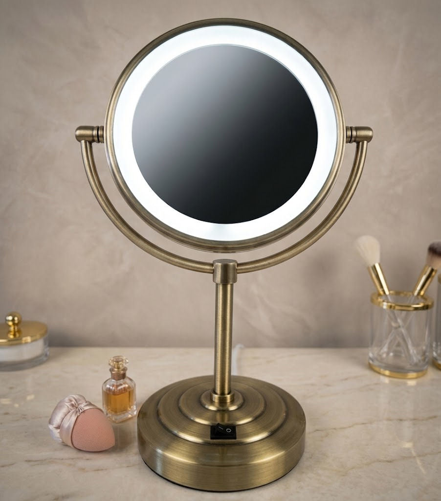 An illuminated vanity mirror with a round LED ring and dual-angle stand, shown on a marble countertop with cosmetic items in a bathroom setting.